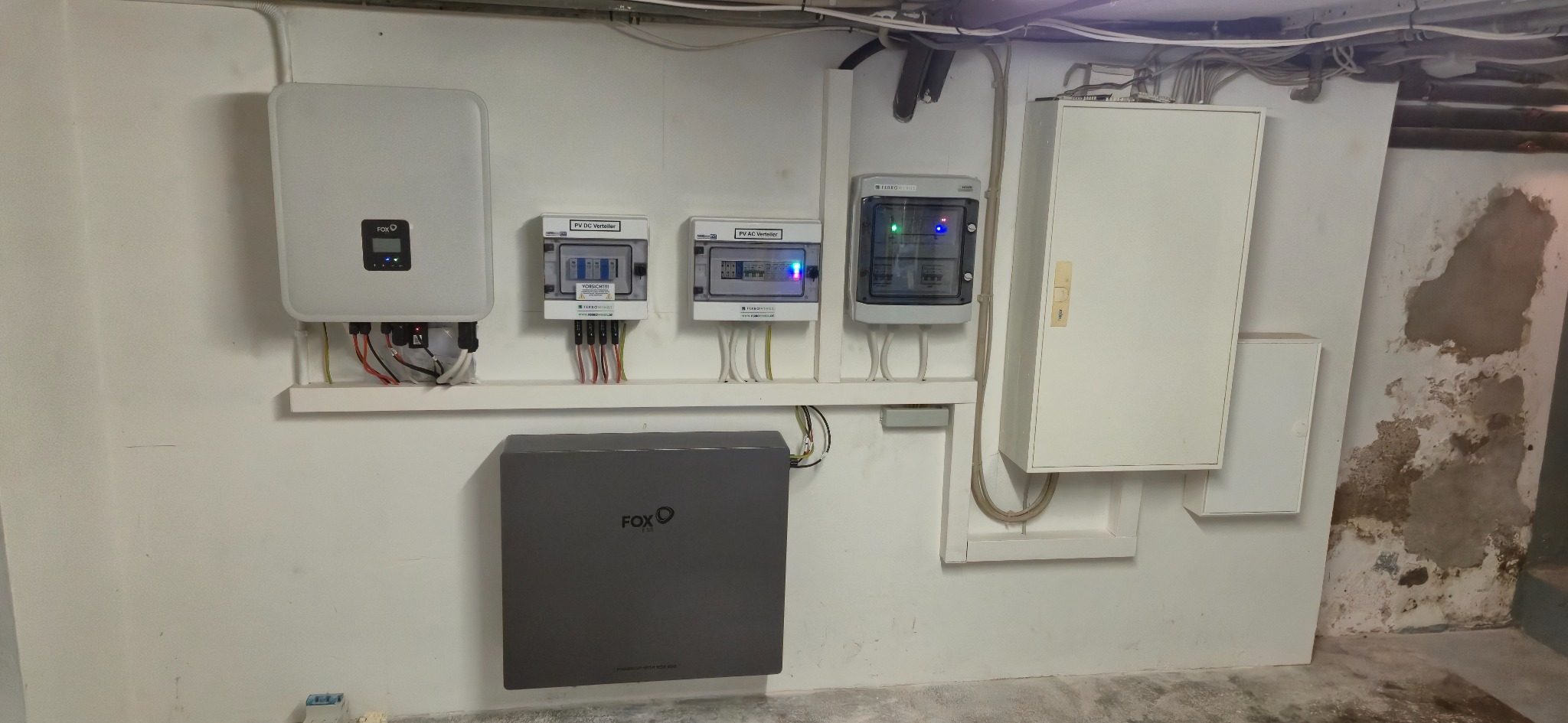 Kompleksowa instalacja: falownik, rozdzielnice AC/DC i magazyn energii FOX na białej ścianie z widocznymi przewodami, w pomieszczeniu o surowym wykończeniu.