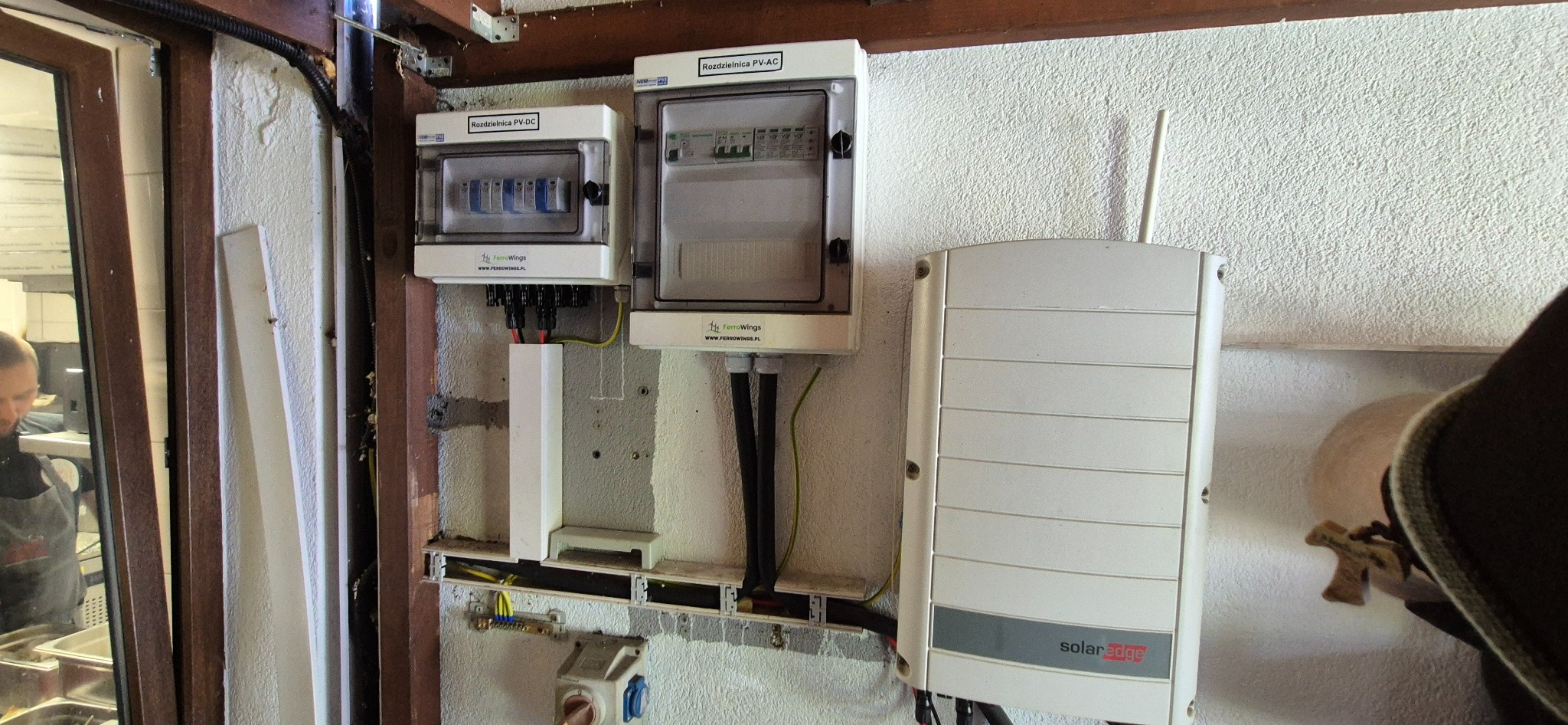 Rozdzielnice PV-DC i PV-AC oraz falownik SolarEdge zamontowane na ścianie z widocznymi przewodami. Elementy instalacji fotowoltaicznej wewnątrz budynku.