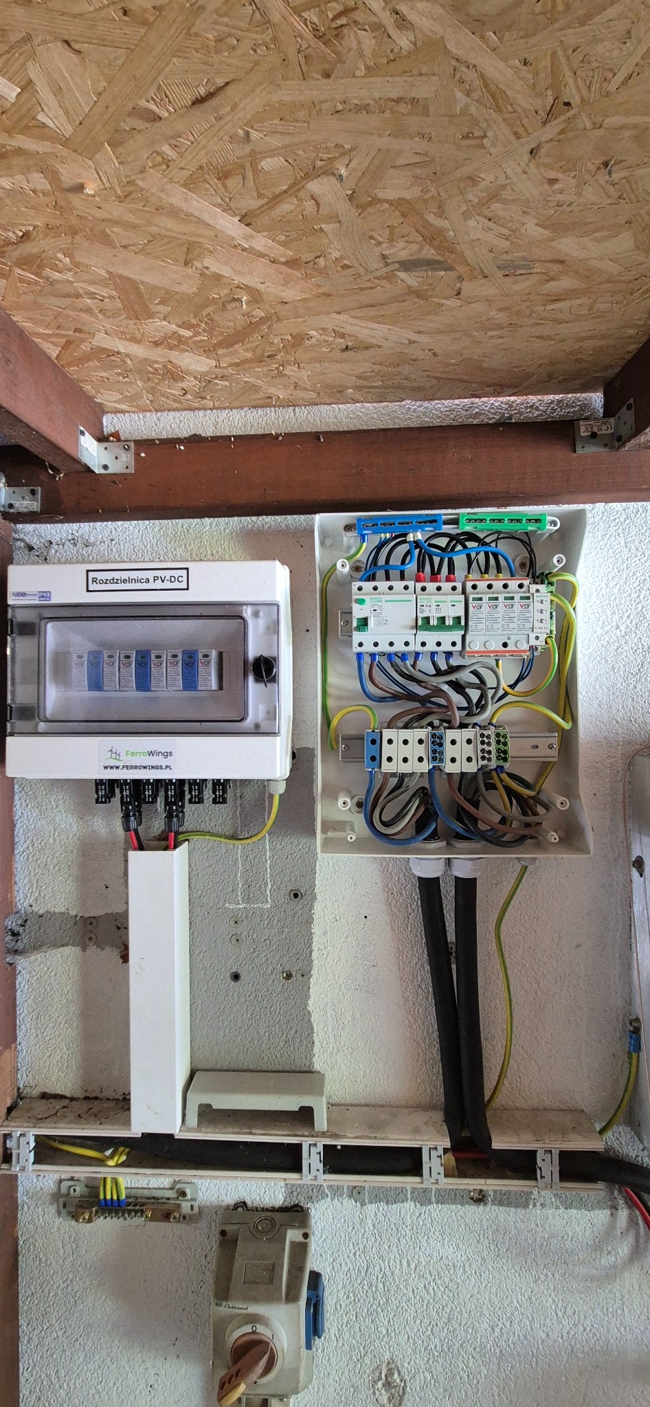 Rozdzielnica PV-DC marki FerroWings z bezpiecznikami i okablowaniem, zamontowana na szarej ścianie wewnątrz budynku z drewnianym stropem.