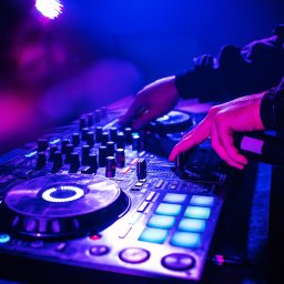 DJ Sławek - DJ miksuje muzykę na profesjonalnym sprzęcie w niebiesko-fioletowym oświetleniu. Widoczne dłonie na mikserze, suwaki, pokrętła i podświetlane przyciski.