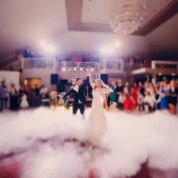 Para młoda tańczy pierwszy taniec w chmurze dymu na tle gości i podświetlonej sceny z nagłośnieniem.