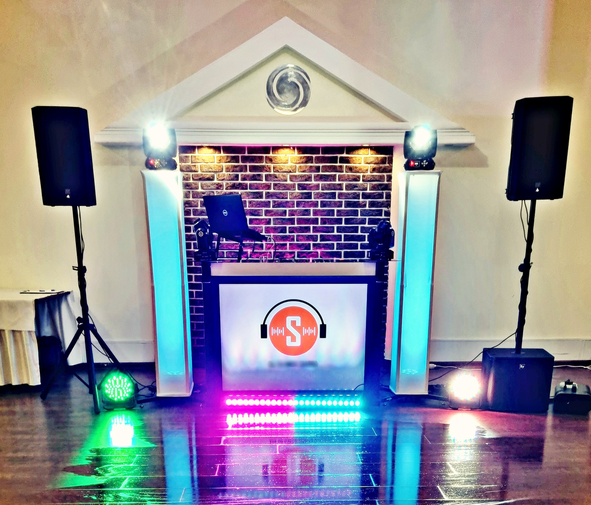 Scena DJ-a z laptopem, reflektorami i głośnikami, podświetlona na fioletowo i zielono, z ceglanym tłem i dekoracyjnym elementem architektonicznym.