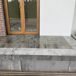 Nowoczesne wejście do domu z kostki brukowej w odcieniach szarości i grafitu, z niskim murkiem. Widoczne drzwi wejściowe i fragment elewacji z cegły.