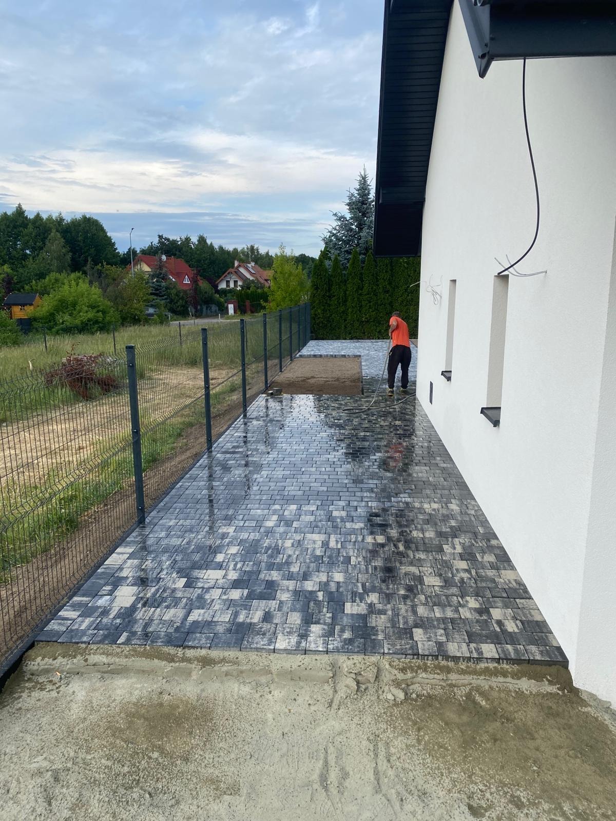 Ułożona, mokra kostka brukowa w odcieniach szarości przy białym budynku. Osoba w pomarańczowej koszulce czyści powierzchnię wodą. Widoczny fragment ogrodzenia panelowego.