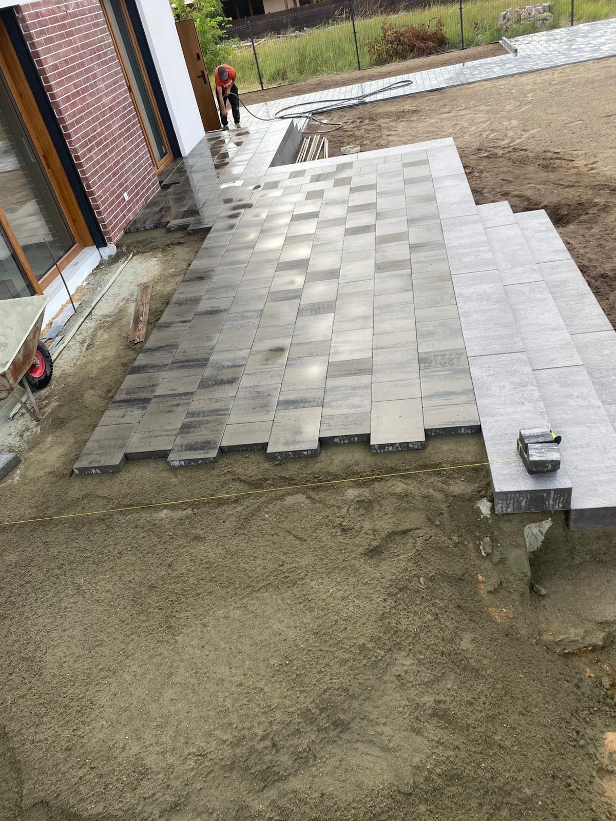 Układanie szarej kostki brukowej na posesji, widoczny fragment tarasu i schodów. W tle dom z ceglaną elewacją i pracownik z wężem. Częściowo ułożona nawierzchnia.
