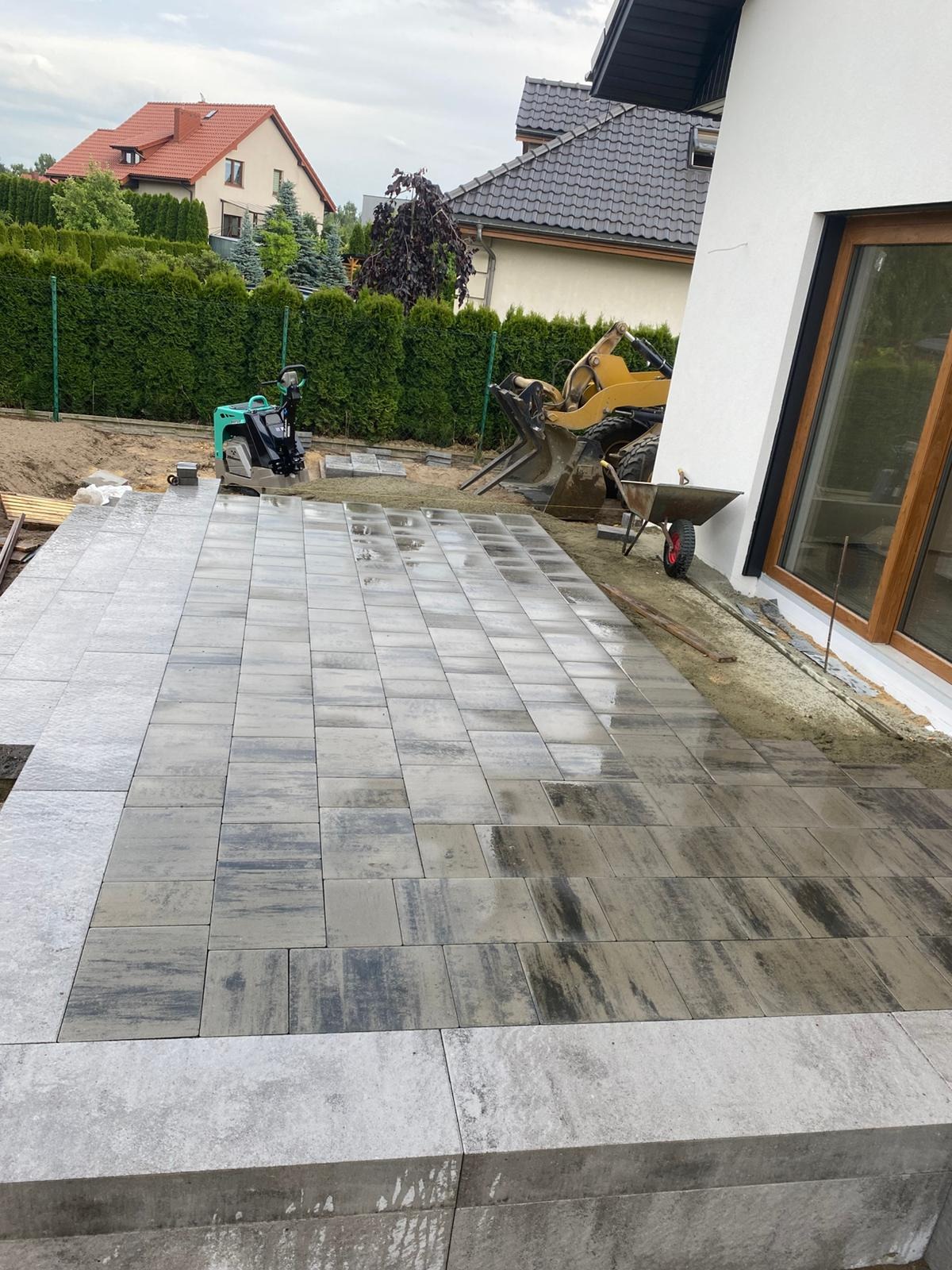 Świeżo ułożona, wilgotna kostka brukowa w odcieniach szarości, z widoczną ubijarką i minikoparką w tle, sugerująca etap prac wykończeniowych na posesji.