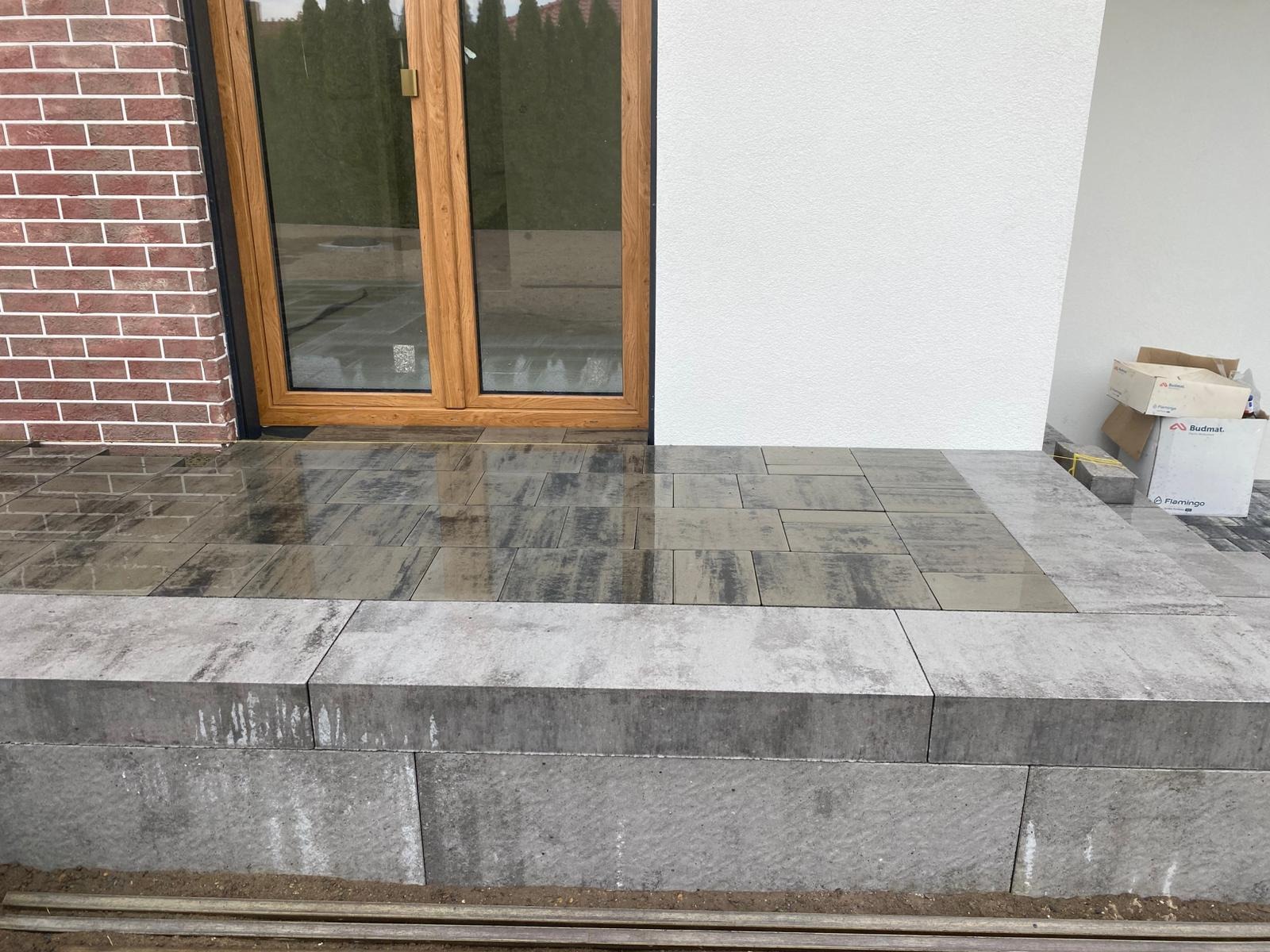 Nowoczesne wejście do domu z kostki brukowej w odcieniach szarości i grafitu, z niskim murkiem. Widoczne drzwi wejściowe i fragment elewacji z cegły.