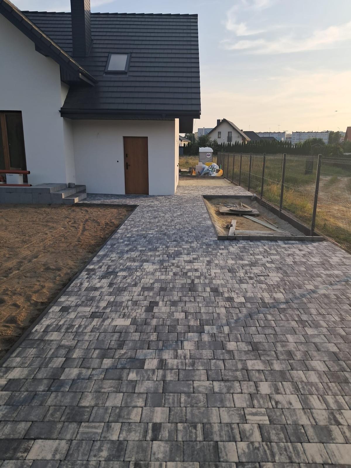 Ułożona kostka brukowa w szaro-białym odcieniu tworzy ścieżkę przed domem. Widoczny fragment trawnika i ogrodzenia. Ujęcie z niskiej perspektywy.