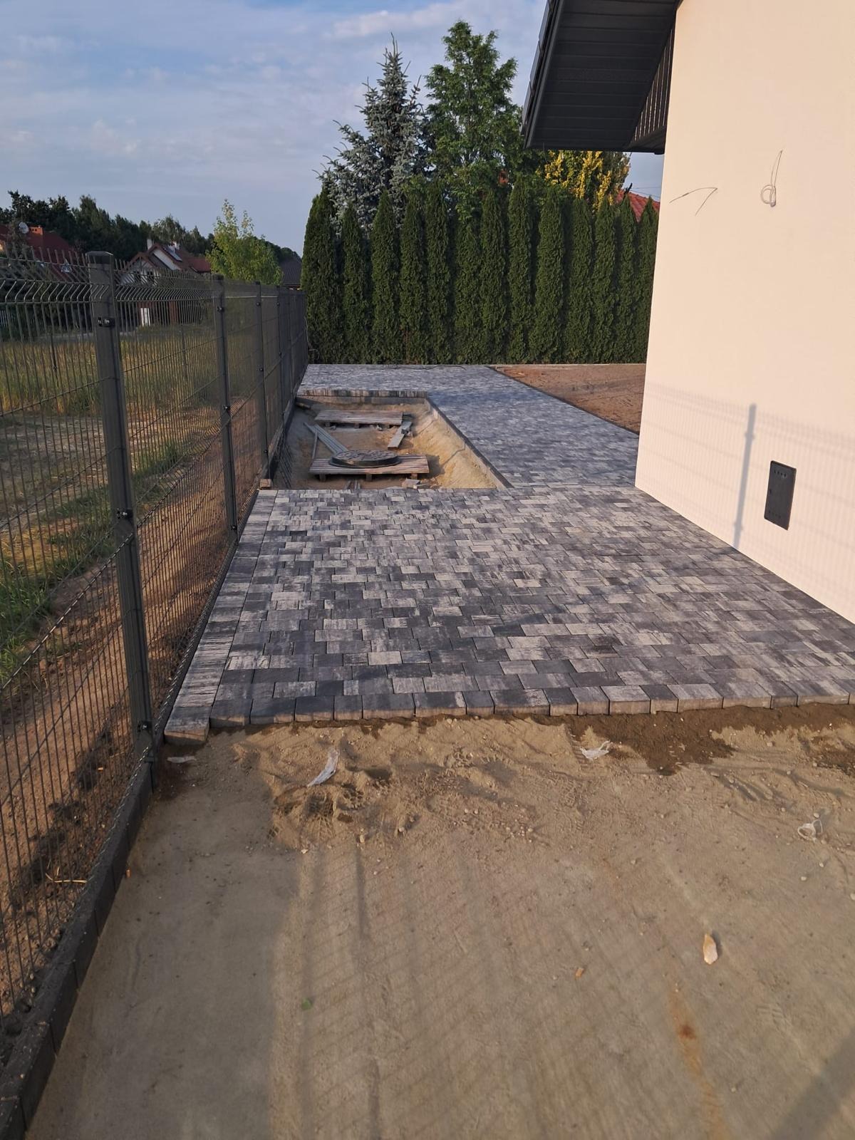 Częściowo ułożona, szara kostka brukowa na podwórku z widocznym wykopem. W tle zielony żywopłot i fragment budynku.