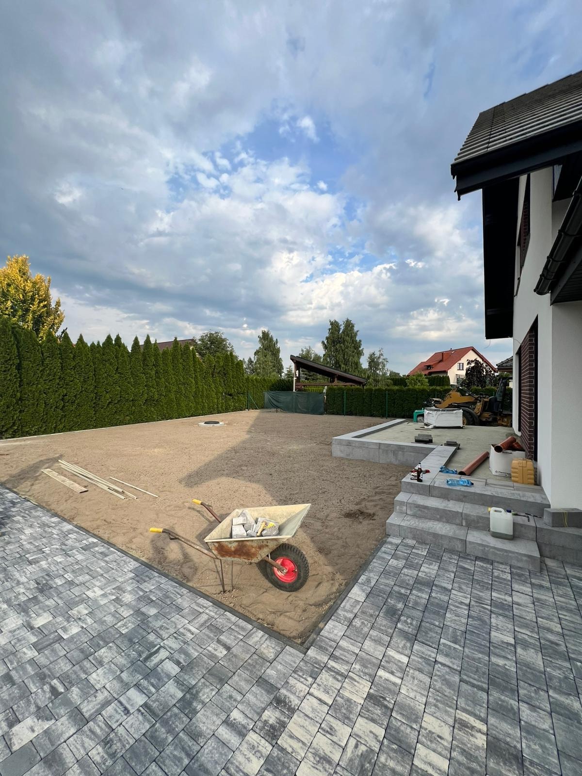 Ułożona kostka brukowa w szaro-białej tonacji, taczka z kostką, przygotowany teren pod dalsze prace, dom z jasną elewacją i ciemnym dachem w tle.