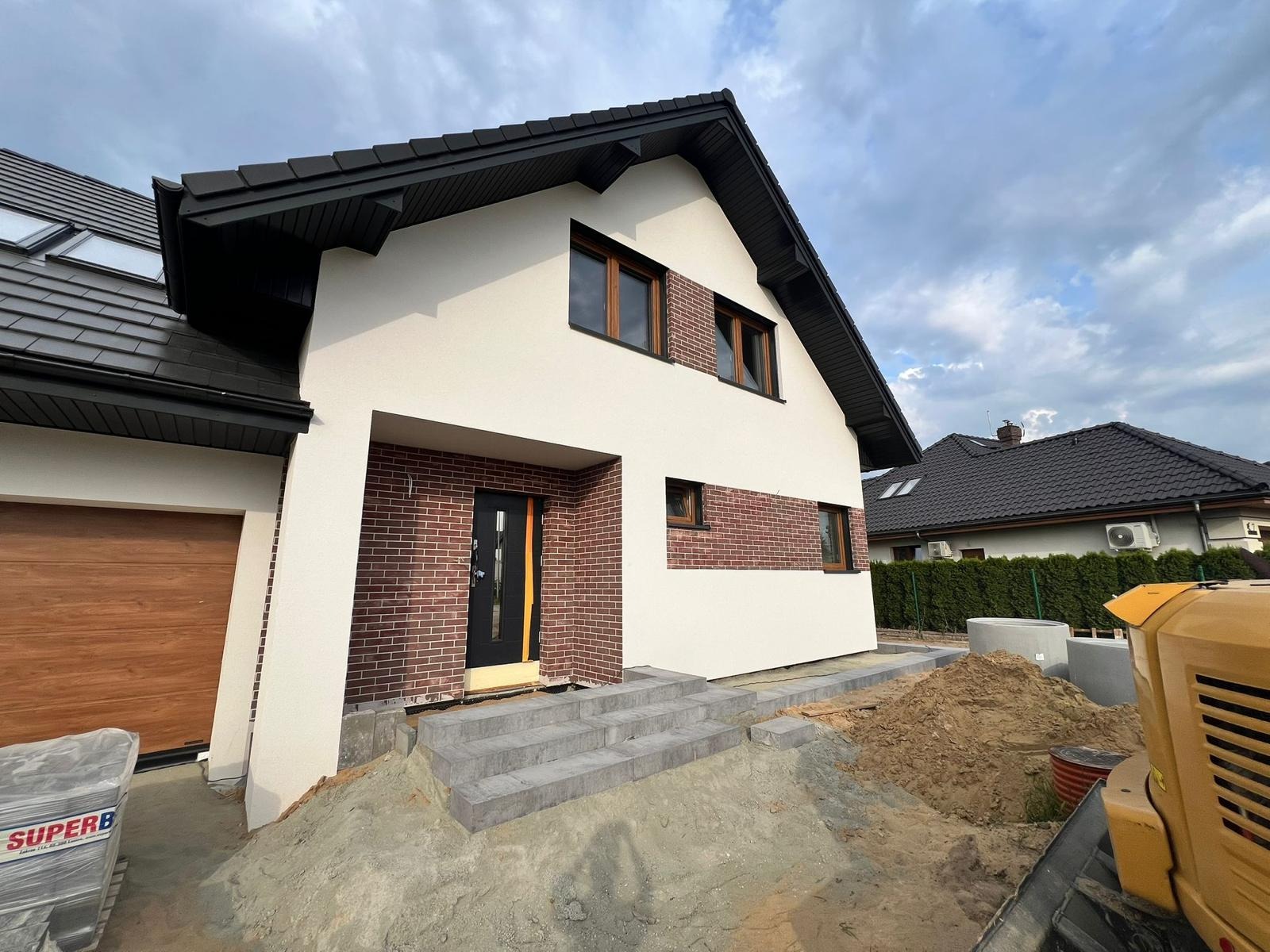 Nowoczesny dom z elewacją w bieli i cegle, z szarymi schodami z kostki brukowej prowadzącymi do drzwi wejściowych. W tle hałda ziemi i fragment maszyny budowlanej.