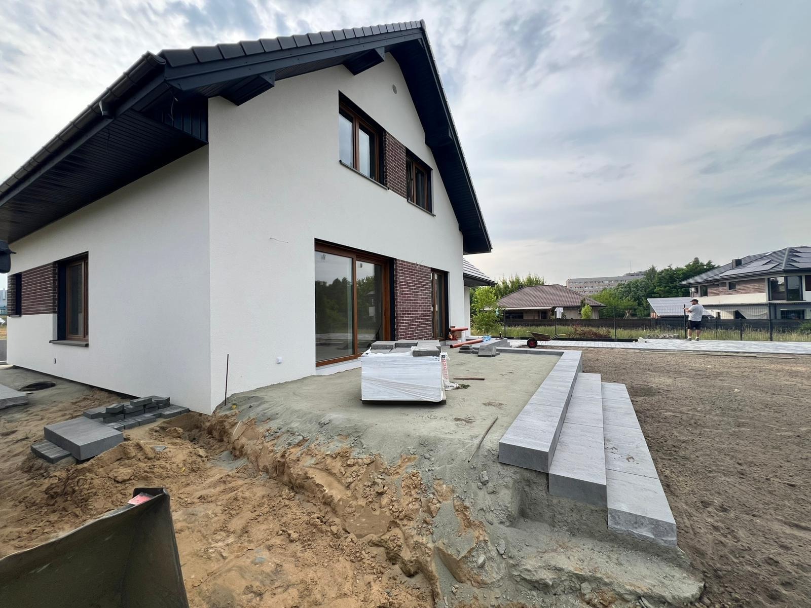 Budowa tarasu z kostki brukowej przy nowoczesnym domu. Widoczne ułożone schody i przygotowany teren pod taras. W tle inne budynki i pochmurne niebo.