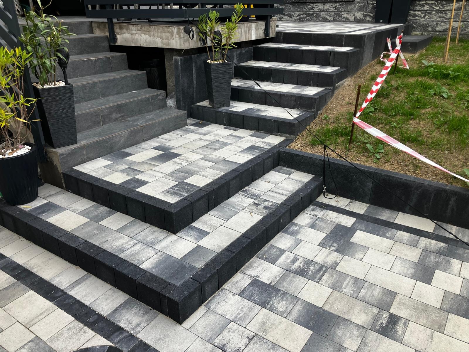 Schody zewnętrzne wyłożone kostką brukową w odcieniach szarości i czerni, z donicami z roślinami po bokach. Widoczne taśmy ostrzegawcze.