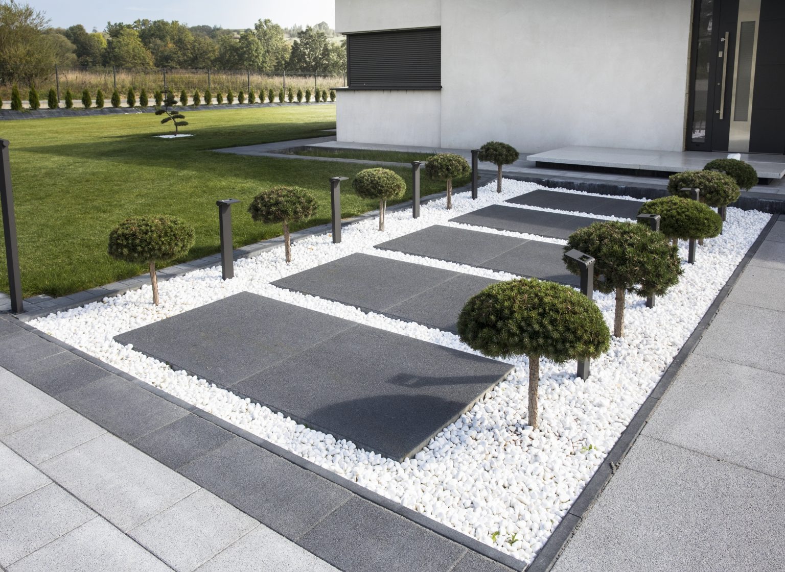 Nowoczesna ścieżka z szarych płyt, białego żwiru i kulistych drzewek prowadzi do domu z minimalistyczną elewacją i czarnymi akcentami.