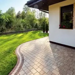 Kremowa kostka brukowa ułożona w łagodny łuk przy budynku z oknem i zielonym trawnikiem. Dekoracyjne obrzeże z ciemniejszej kostki. Dwa tuje w donicach.