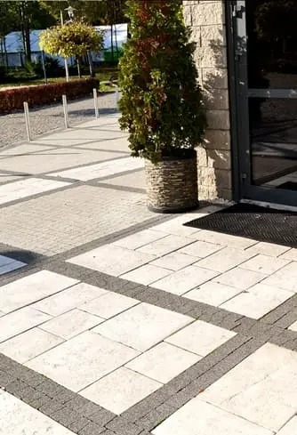 Jasna kostka brukowa ułożona w geometryczny wzór z ciemniejszym obramowaniem. Donica z rośliną przy wejściu do budynku. Widok z góry, lekki cień.