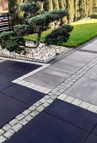 Nowoczesna nawierzchnia z kostki brukowej w odcieniach szarości i czerni, z dekoracyjnym obramowaniem z kostki granitowej i elementami zieleni w tle.