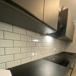 Axel Styl Oleksii Hrybkov - Białe, błyszczące płytki na ścianie nad blatem kuchennym, z czarną fugą i ciemnymi szafkami wiszącymi, oświetlone światłem z okapu. Nowoczesny design.