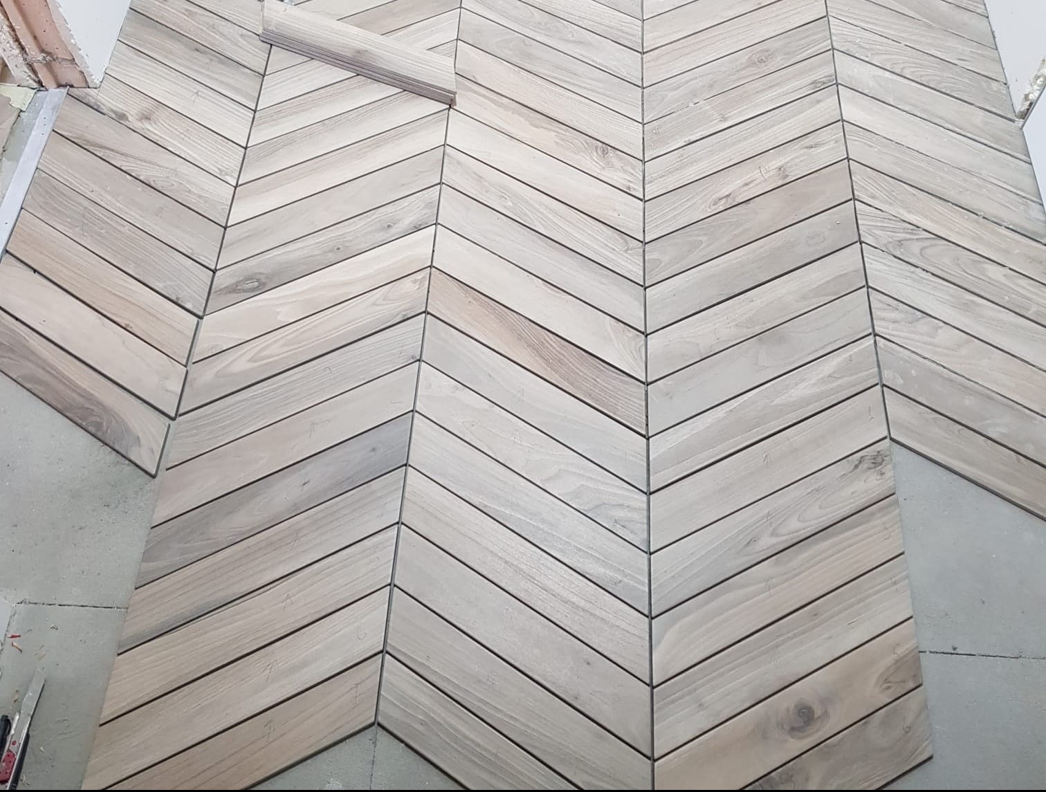 Układanie jasnych, drewnianych paneli podłogowych w jodełkę na surowym podłożu. Widoczne niedokończone krawędzie i pojedyncze panele gotowe do montażu.