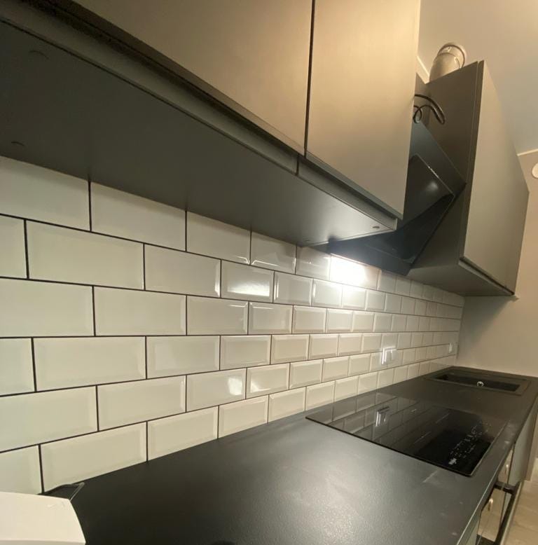 Białe, błyszczące płytki na ścianie nad blatem kuchennym, z czarną fugą i ciemnymi szafkami wiszącymi, oświetlone światłem z okapu. Nowoczesny design.