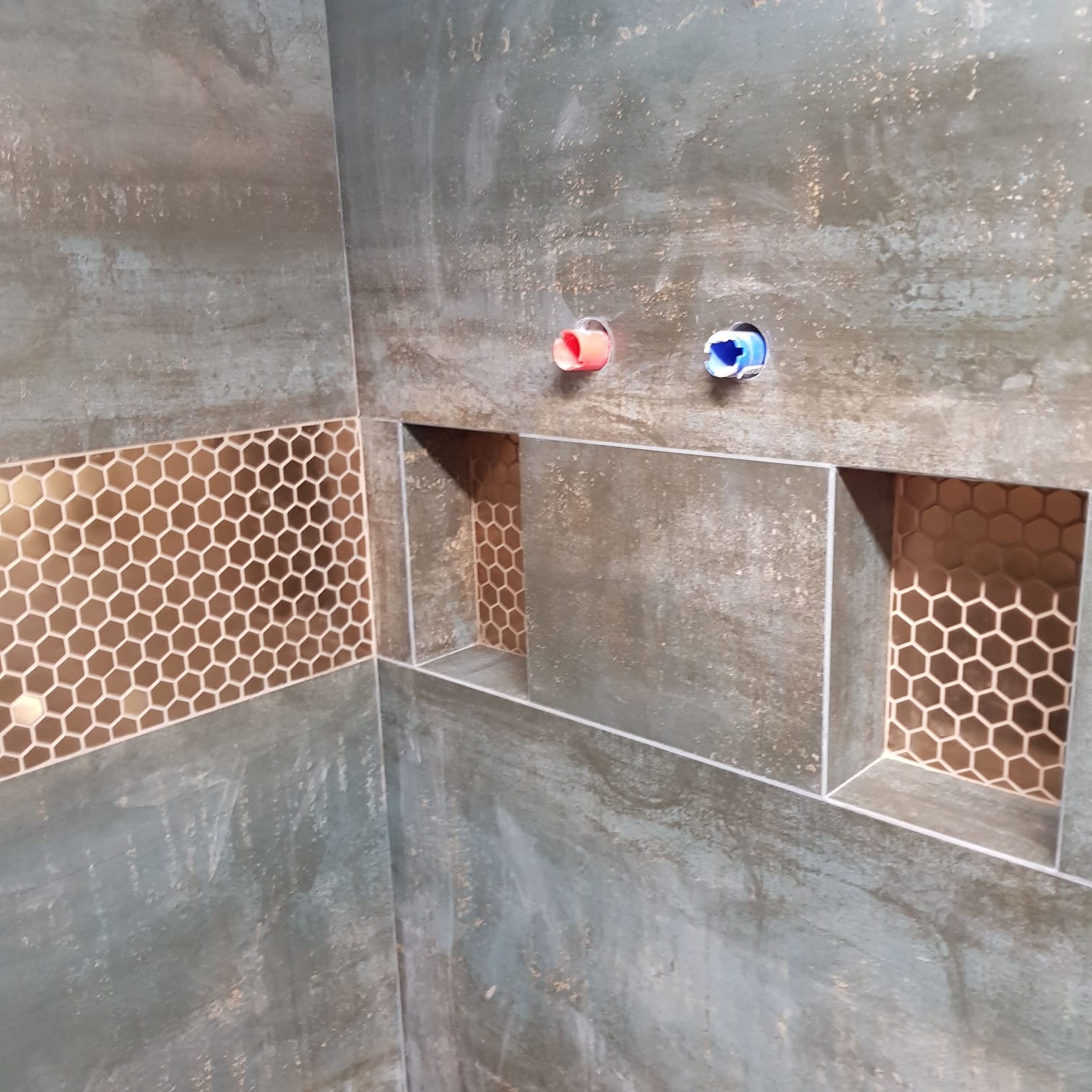 Wykończona ściana łazienki z szarozielonymi płytkami, wnękami wyłożonymi mozaiką w kształcie plastra miodu i widocznymi przyłączami wodnymi (ciepła i zimna).