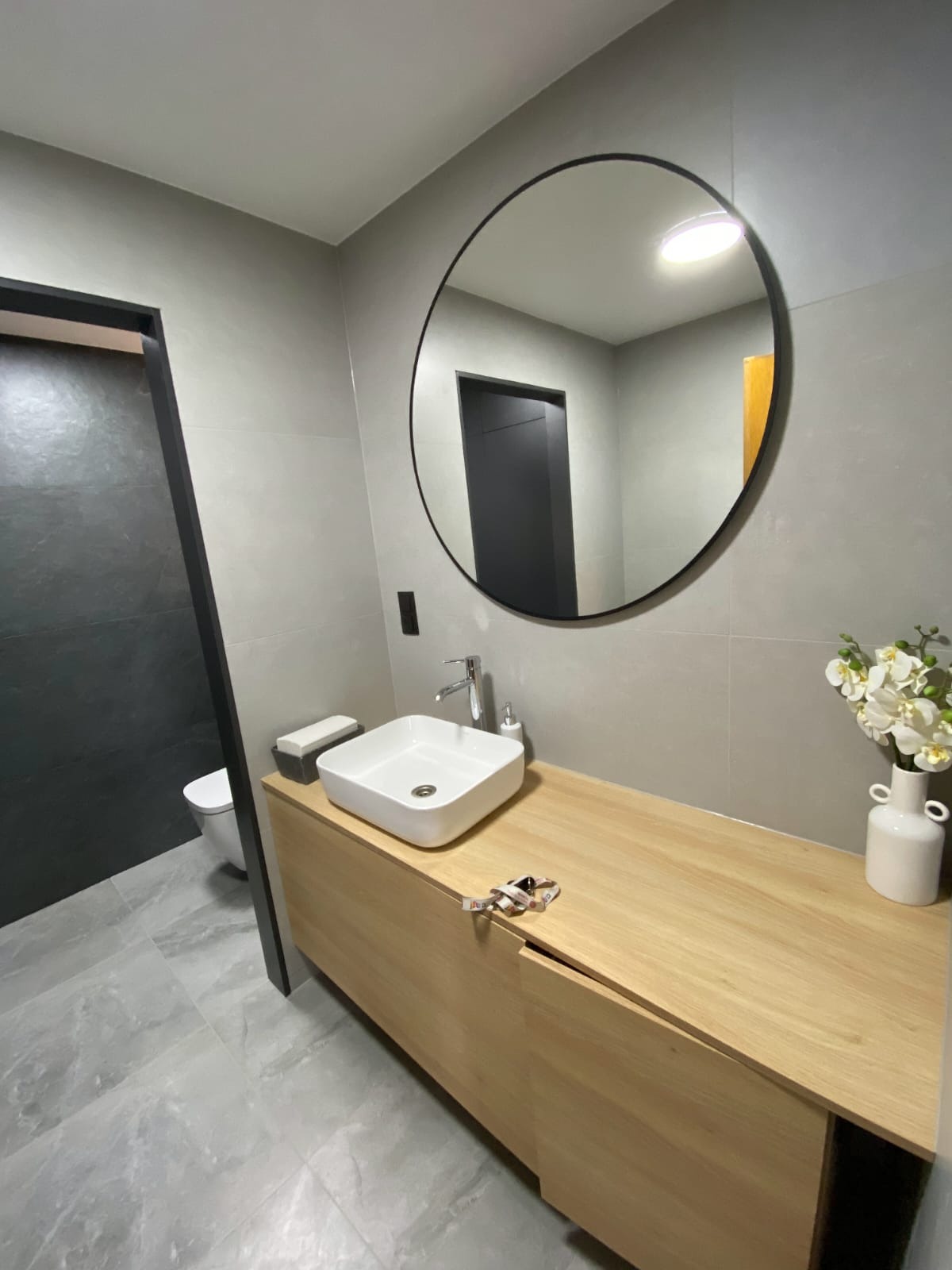Nowoczesna łazienka z okrągłym lustrem, jasną szafką pod umywalką, kwadratową umywalką i szarą glazurą. Minimalistyczny design z akcentem kwiatowym.