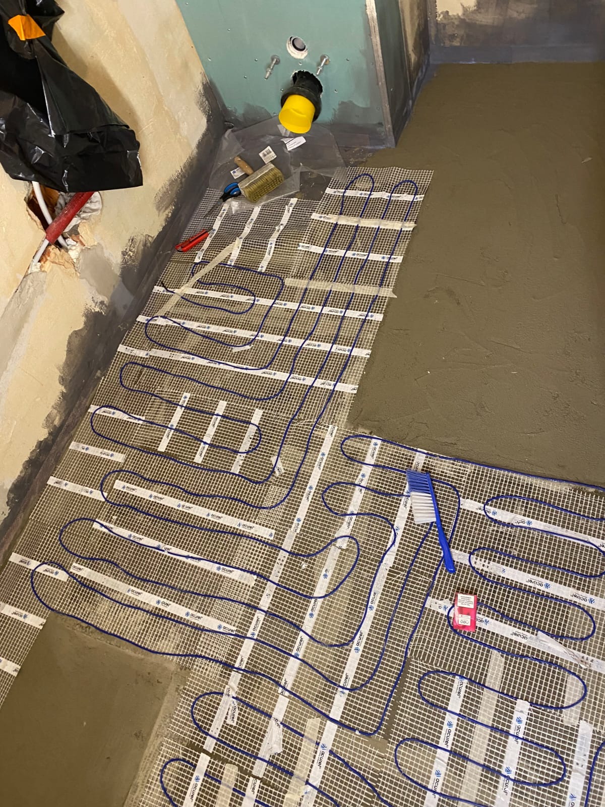 Montaż ogrzewania podłogowego w łazience: niebieskie kable na siatce, częściowo zalane wylewką. Widoczne narzędzia i instalacja sanitarna.