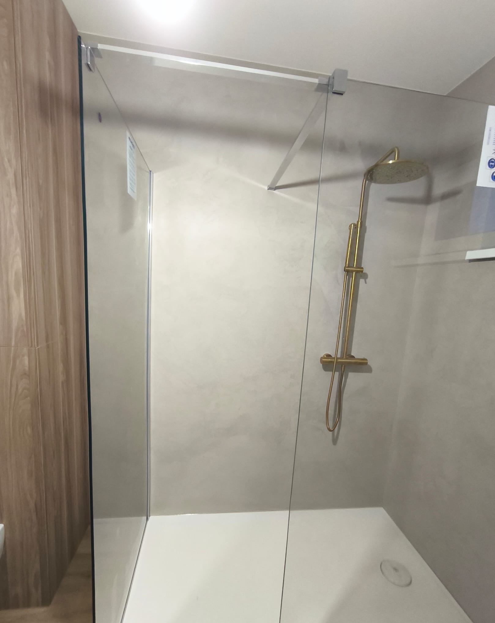 Nowoczesna kabina prysznicowa z transparentnego szkła, złota armatura. Minimalistyczny design łazienki z beżowymi ścianami i drewnopodobną okładziną.