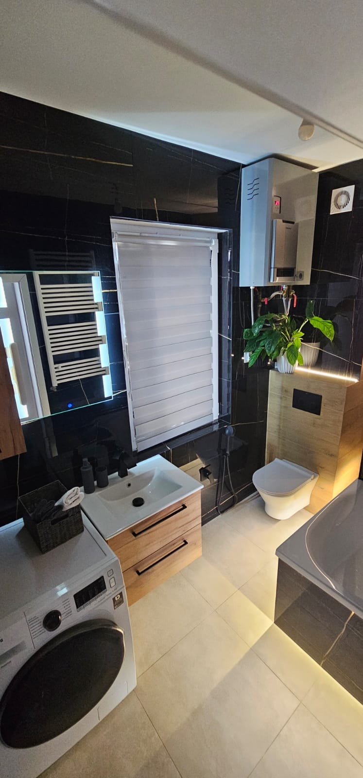 Elegancka łazienka w czerni i drewnie: umywalka z szafką, toaleta z rośliną, pralka, wanna i grzejnik. Czarne płytki z złotymi żyłami, roleta w oknie.