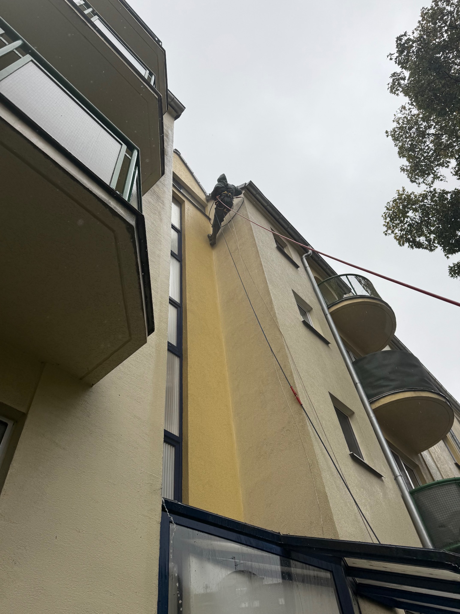 Pracownik na linie zabezpieczającej, podczas inspekcji elewacji budynku wielorodzinnego z balkonami. Ujęcie z dołu, pochmurne niebo w tle.