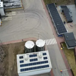 Widok z drona na instalacje fotowoltaiczne na budynkach przemysłowych i wiacie parkingowej oraz zbiorniki. Zauważalne ślady opon na placu.