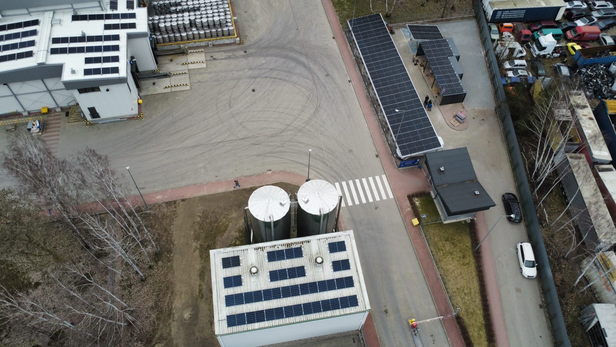 Widok z drona na instalacje fotowoltaiczne na budynkach przemysłowych i wiacie parkingowej oraz zbiorniki. Zauważalne ślady opon na placu.
