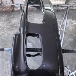 Polakierowany na ciemny metalik zderzak samochodowy, zawieszony na metalowym stojaku w warsztacie, z widocznymi refleksami światła na gładkiej powierzchni.