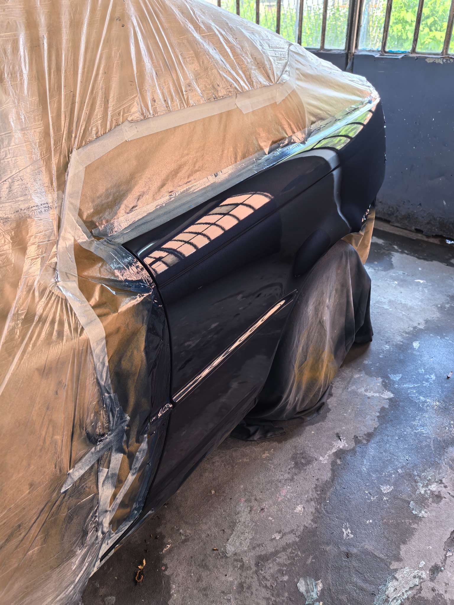 Częściowo zakryty folią i taśmą samochód w trakcie lakierowania, z widocznym fragmentem świeżo polakierowanego, czarnego błotnika. Widok na warsztat przez okno.