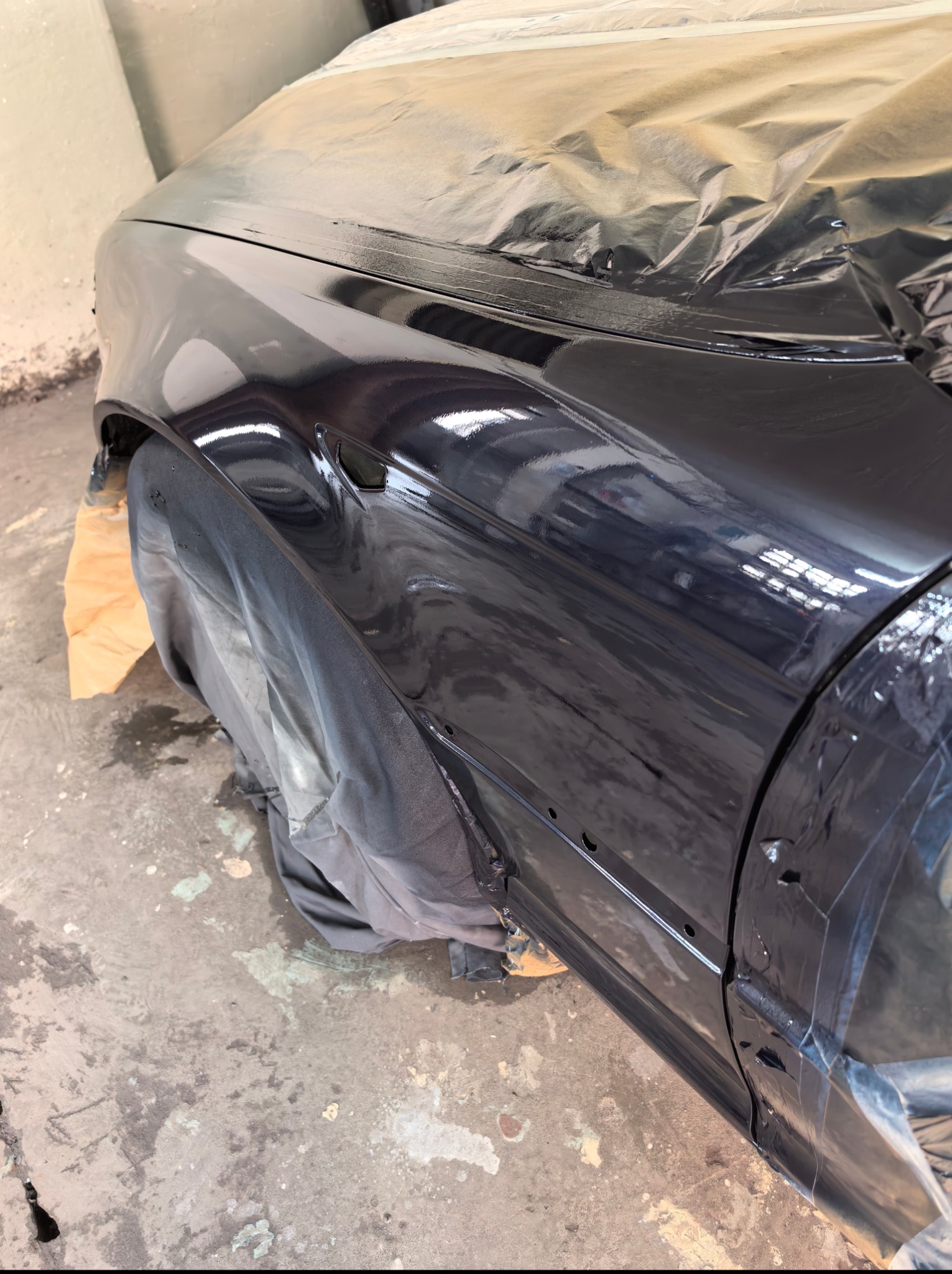 Częściowo zakryty folią, świeżo polakierowany na ciemny kolor fragment karoserii samochodu. Widoczne ślady farby na podłodze warsztatu.