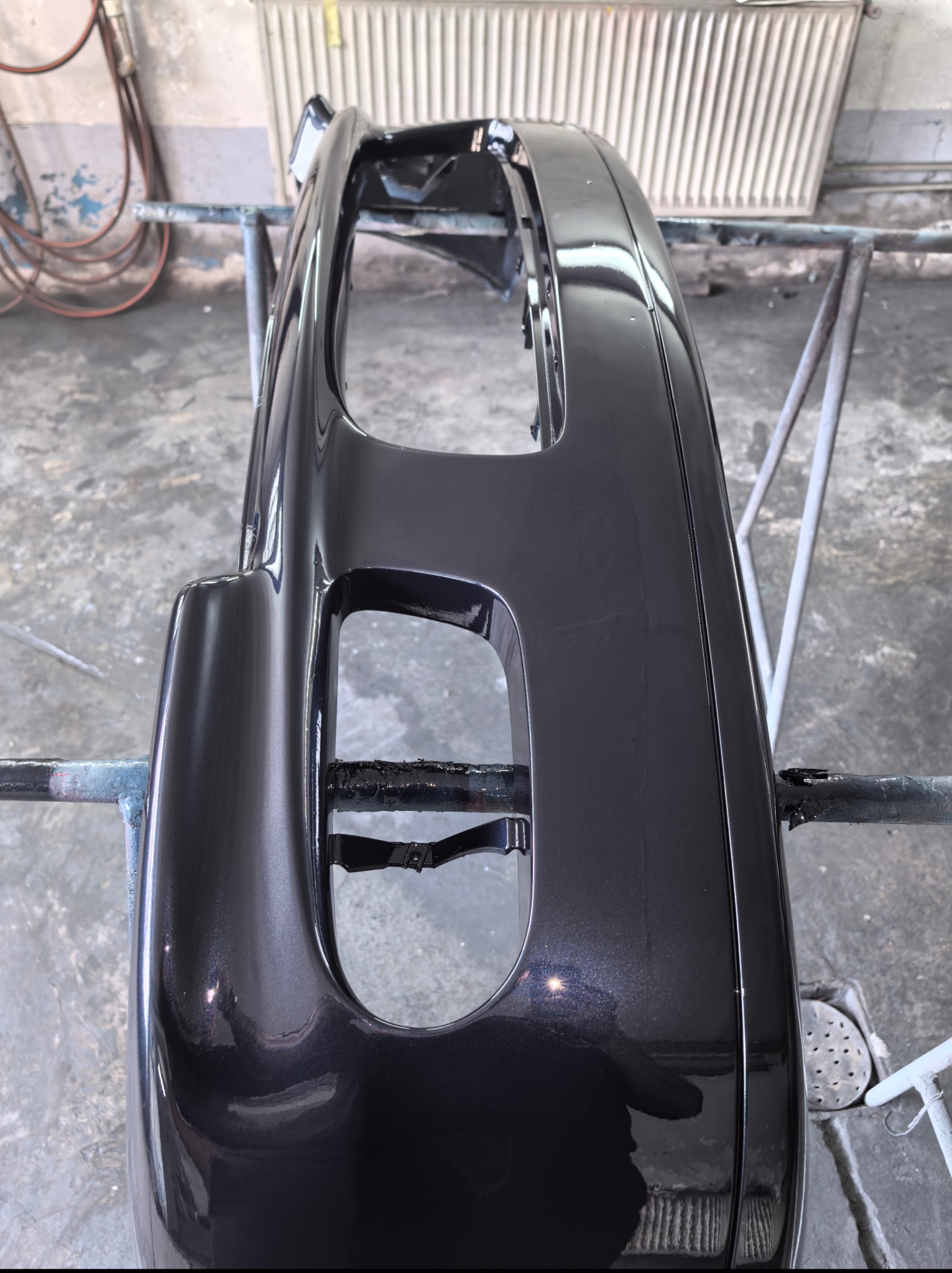 Polakierowany na ciemny metalik zderzak samochodowy, zawieszony na metalowym stojaku w warsztacie, z widocznymi refleksami światła na gładkiej powierzchni.