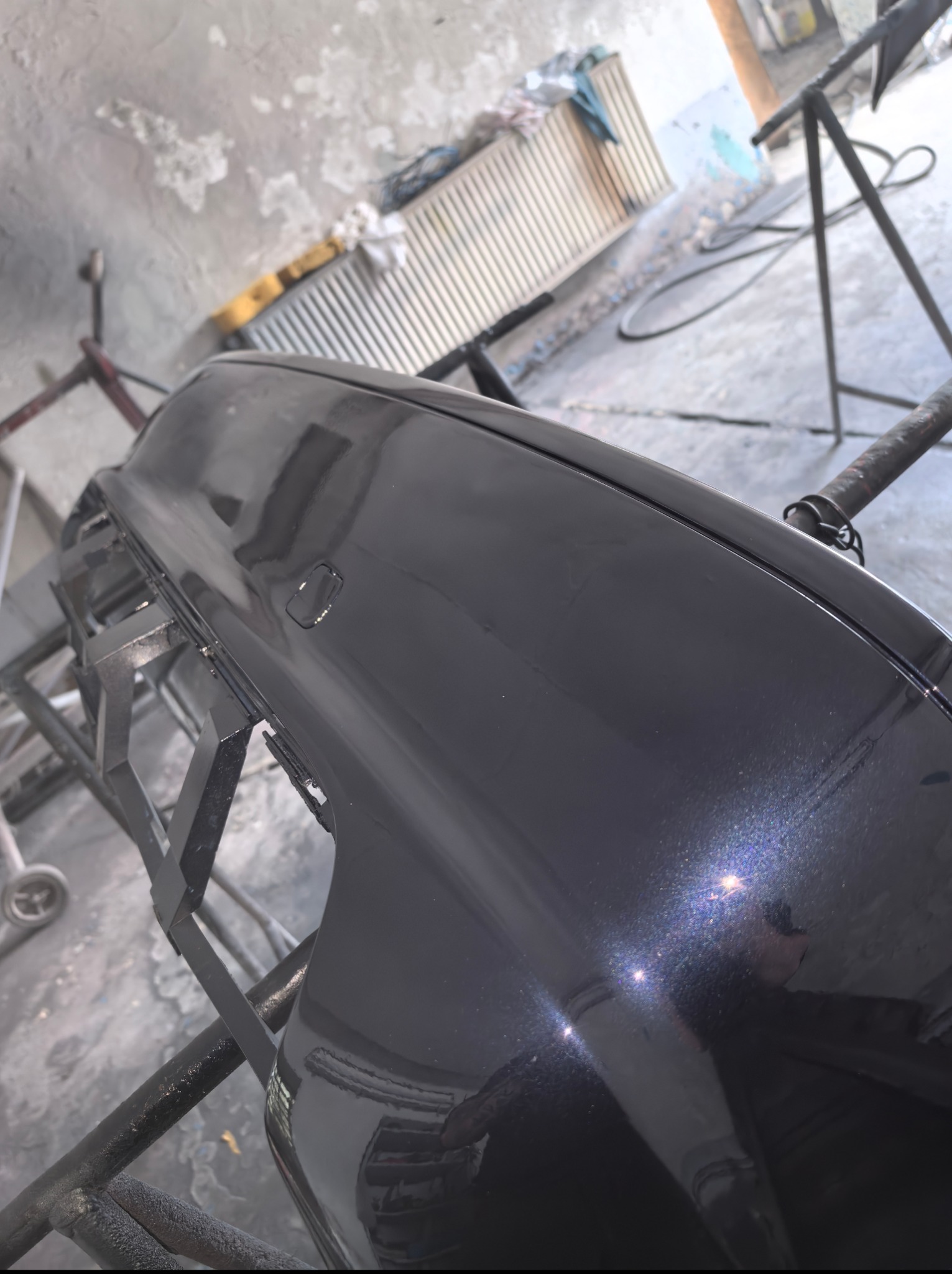 Polakierowany zderzak samochodowy w warsztacie w Mysłowicach. Błyszcząca, ciemna powierzchnia z efektem metalic. Zderzak umieszczony na metalowym stojaku.