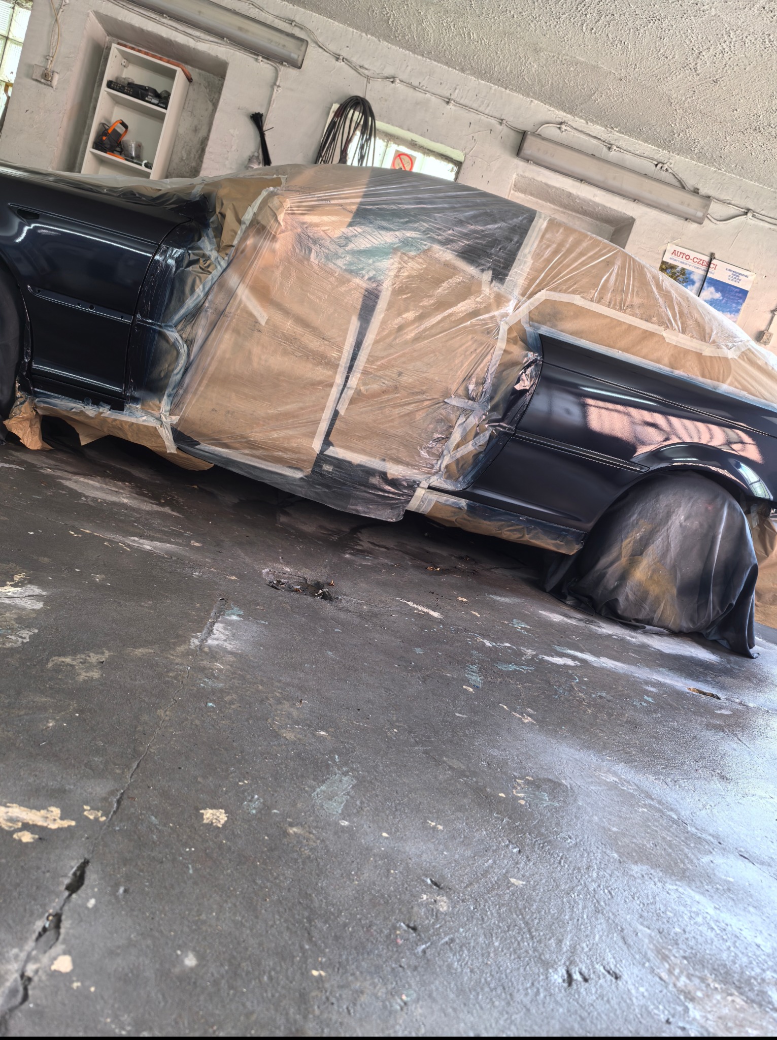 Częściowo zaklejony folią i papierem samochód w trakcie lakierowania na ciemny kolor w warsztacie samochodowym, widok z boku na błotnik i drzwi.