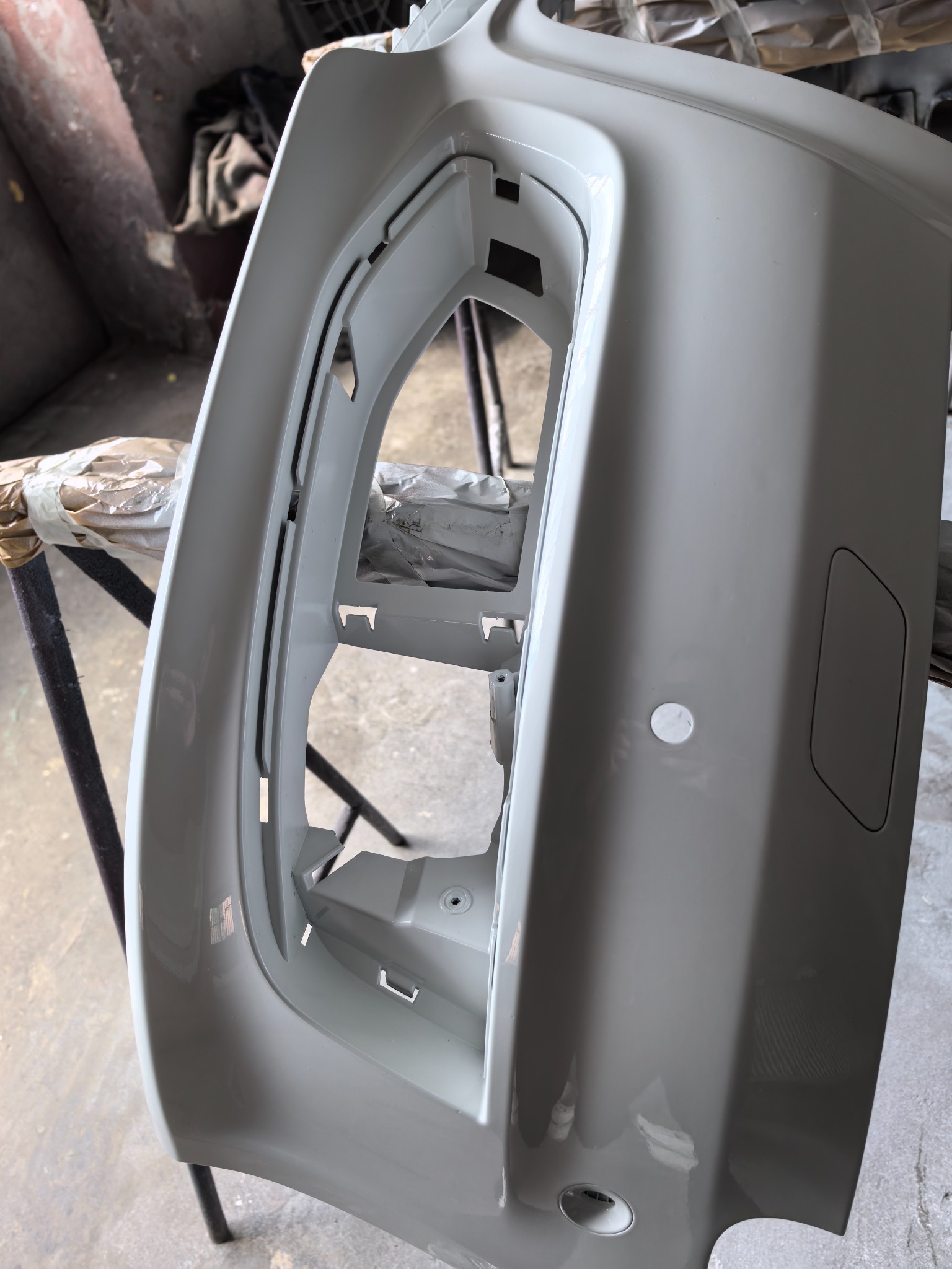 Pomalowany na szaro przedni zderzak samochodowy, umieszczony na stojaku w warsztacie. Widoczne detale i otwory montażowe.