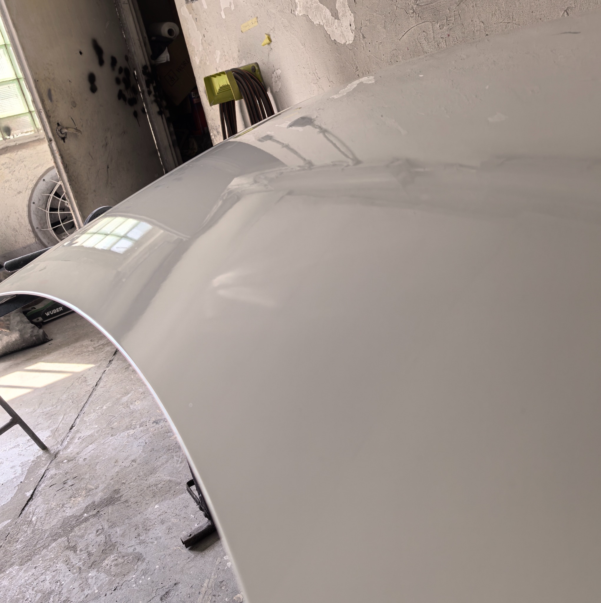 Pomalowana na szaro maska samochodu w warsztacie, widoczne odbicia światła i wentylator w tle, co sugeruje etap suszenia lakieru.