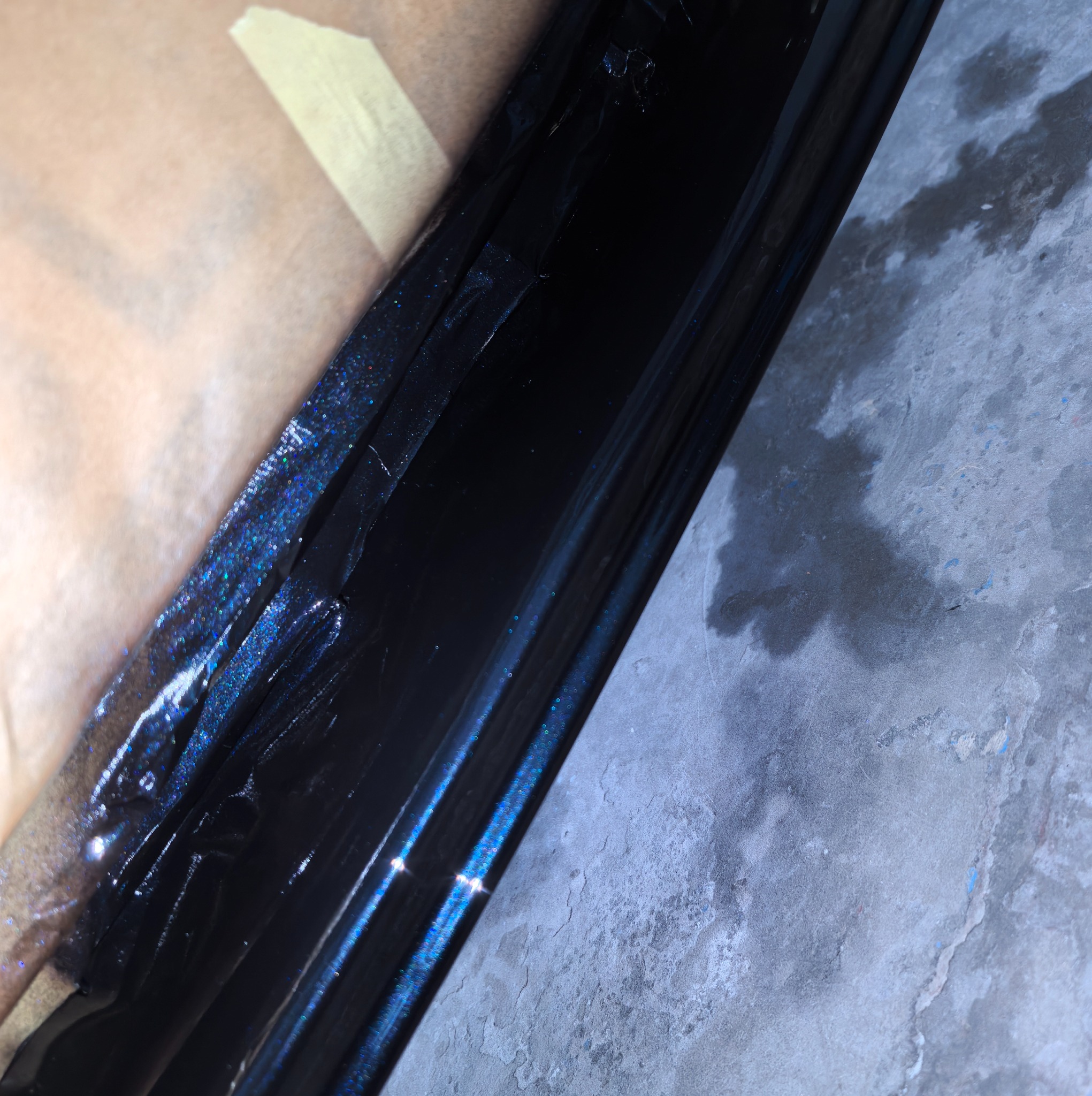 Błyszczący, czarny element karoserii samochodu po lakierowaniu, zabezpieczony taśmą malarską, leżący na betonowej posadzce. Widoczne drobinki brokatu w lakierze.