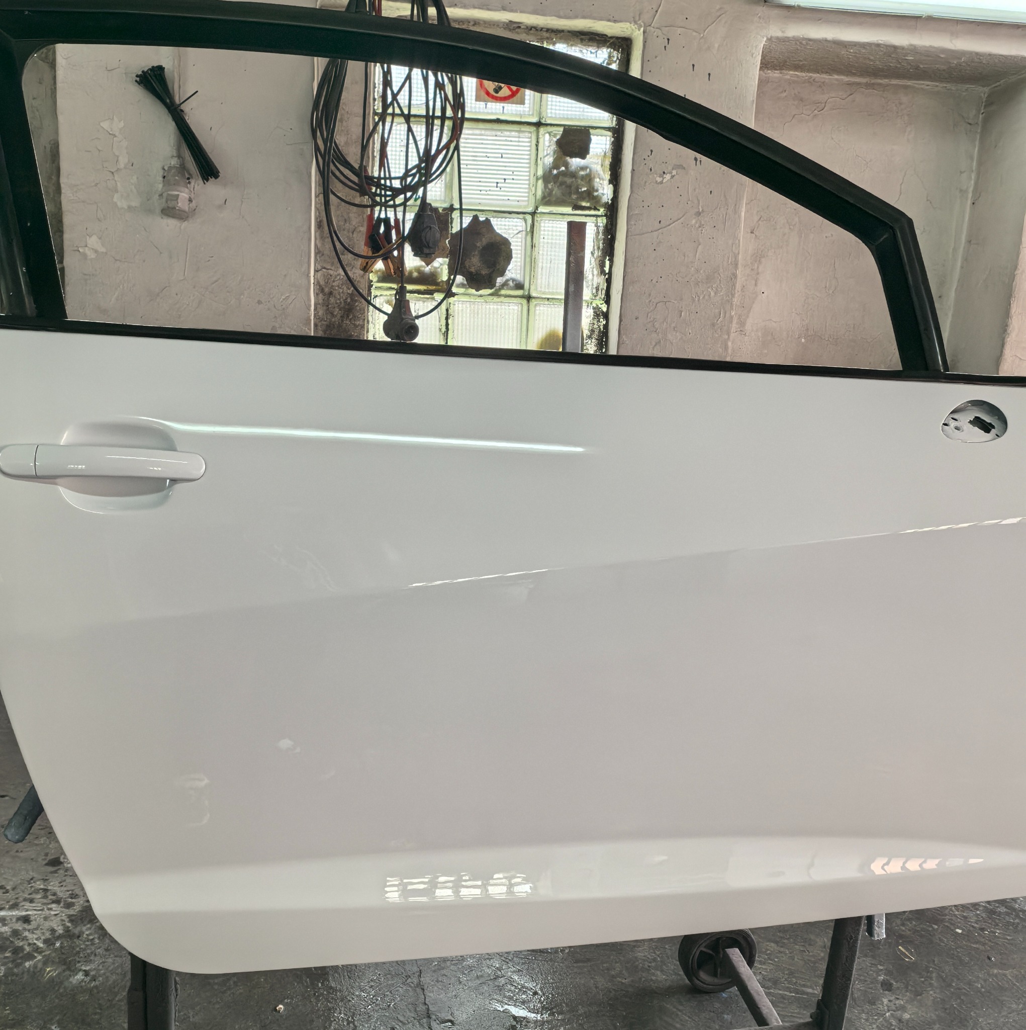 Białe drzwi samochodowe po lakierowaniu, zawieszone w warsztacie. Widoczne refleksy światła, uchwyt i fragment ramy okiennej z zewnątrz.