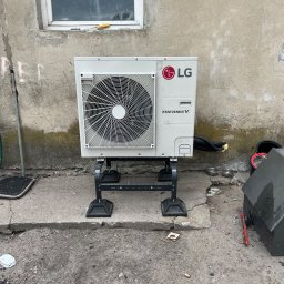 Royal Eco Energy - Biała zewnętrzna jednostka pompy ciepła LG Therma V, ustawiona na metalowym stelażu na tle szarej ściany z oknem. Widoczne elementy instalacji i otoczenia.