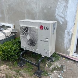 Royal Eco Energy - Biała pompa ciepła LG Therma V, zamontowana na czarnej konstrukcji, stoi przed surową ścianą budynku. Obok widoczne rośliny, taczka z kamieniami i okno.