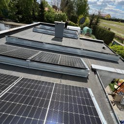 Royal Eco Energy - Instalacja fotowoltaiczna na płaskim dachu budynku jednorodzinnego, widok z góry, panele w metalowych ramach, w tle zieleń drzew i błękitne niebo.