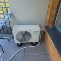 Kompaktowa jednostka zewnętrzna klimatyzacji Kaisai Eco na balkonie, umieszczona na gumowych podkładkach antywibracyjnych, z widoczną białą rurą odprowadzającą skropliny.