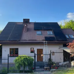 Dom jednorodzinny z częściowo zamontowanymi panelami fotowoltaicznymi na dachu z czerwoną dachówką, drabina oparta o dach, błękitne niebo.