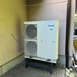 Biała pompa ciepła Panasonic R32 na zewnątrz budynku, ustawiona na czarnym stelażu, z widocznymi elementami instalacji. Obok rynna spustowa.