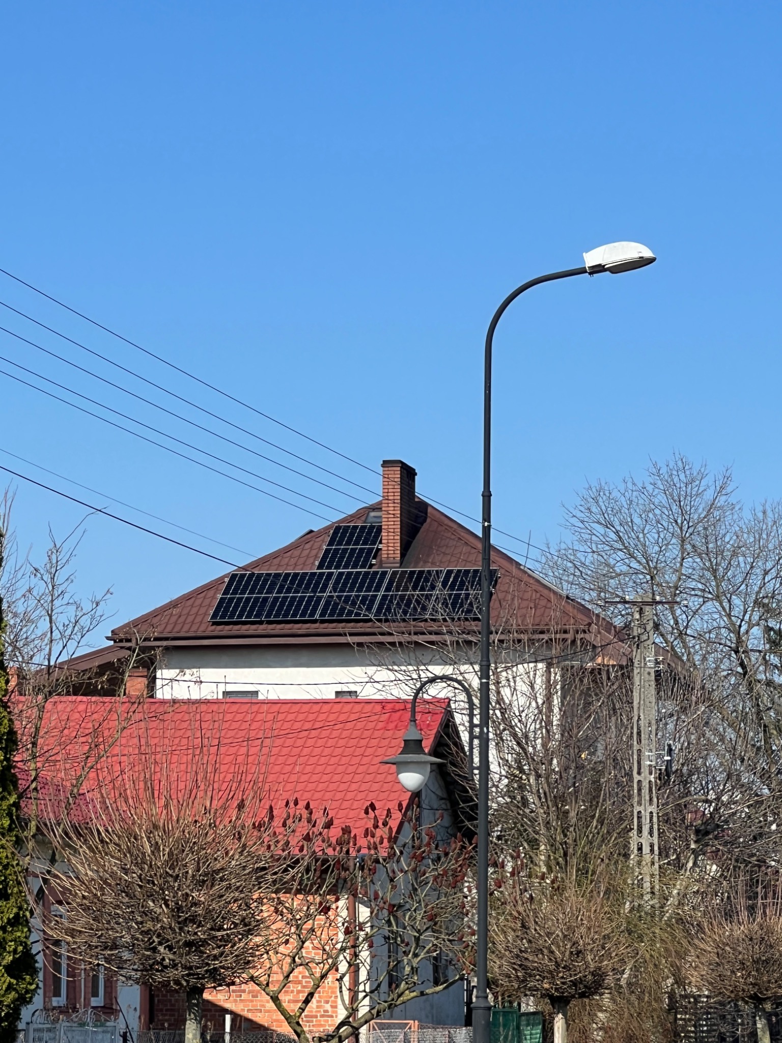 Dom z brązowym dachem i panelami słonecznymi na dachu, widoczny zza drzew i latarni ulicznej na tle błękitnego nieba w słoneczny dzień.