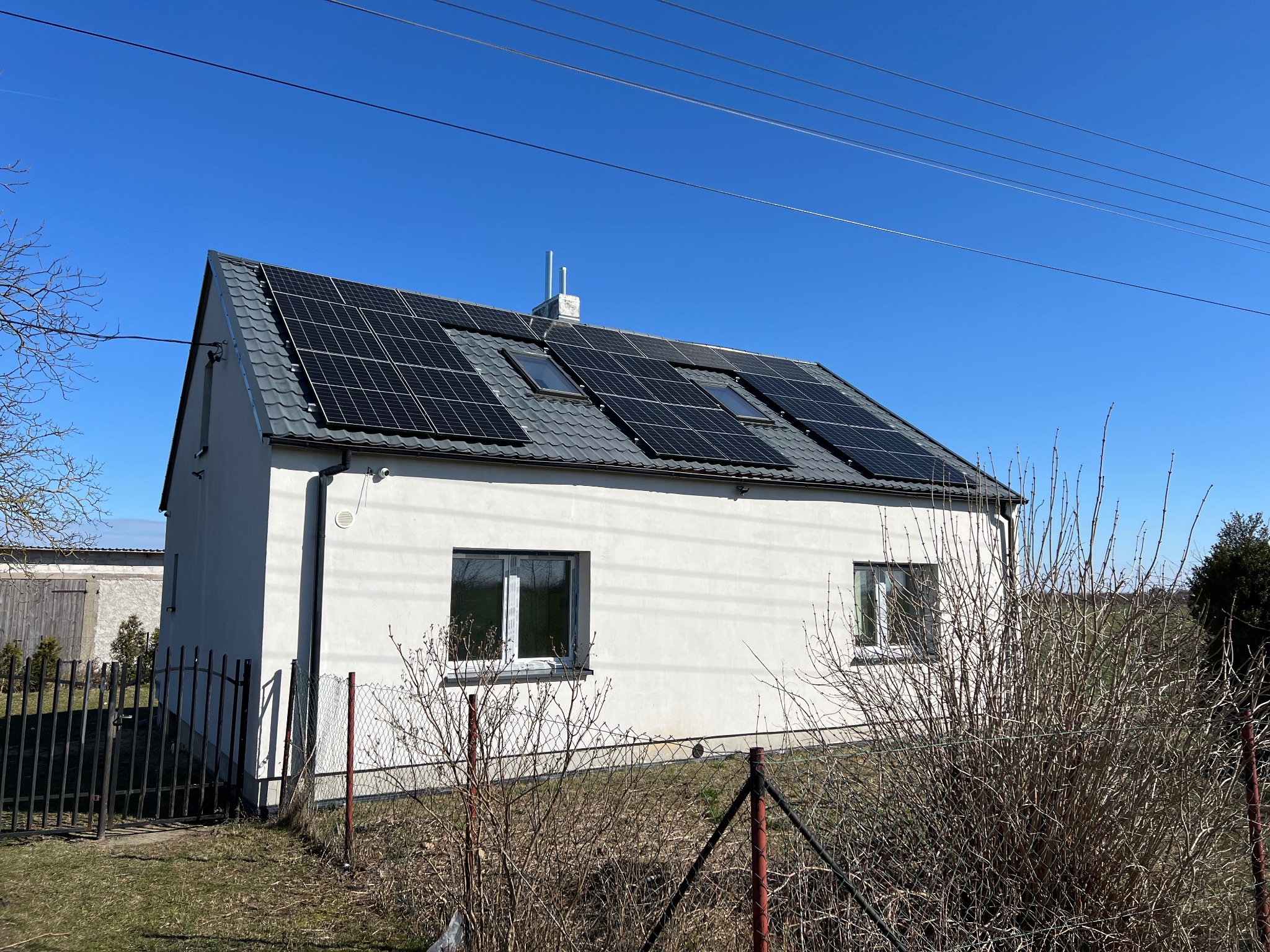 Biały dom z panelami fotowoltaicznymi na szarym dachu, widoczne okna dachowe. Błękitne niebo w tle, ogrodzenie i roślinność na pierwszym planie.